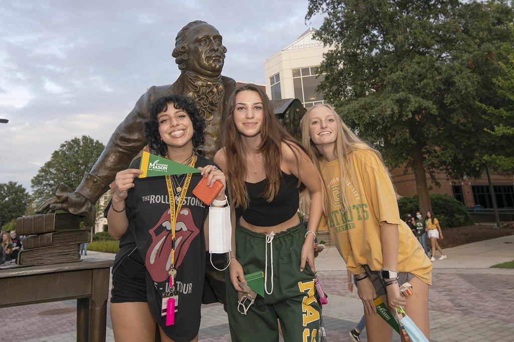 Three young women in Mason gear posing in front of the George Mason statue