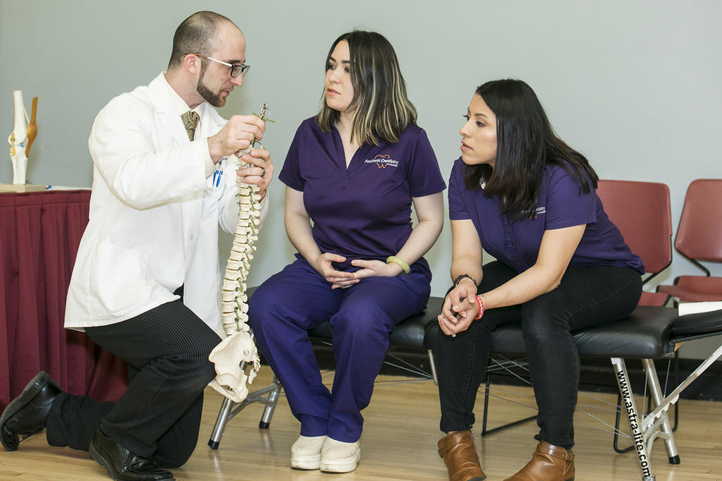 Man in white coat holding a model of the spine talking with two women in purple scrubs